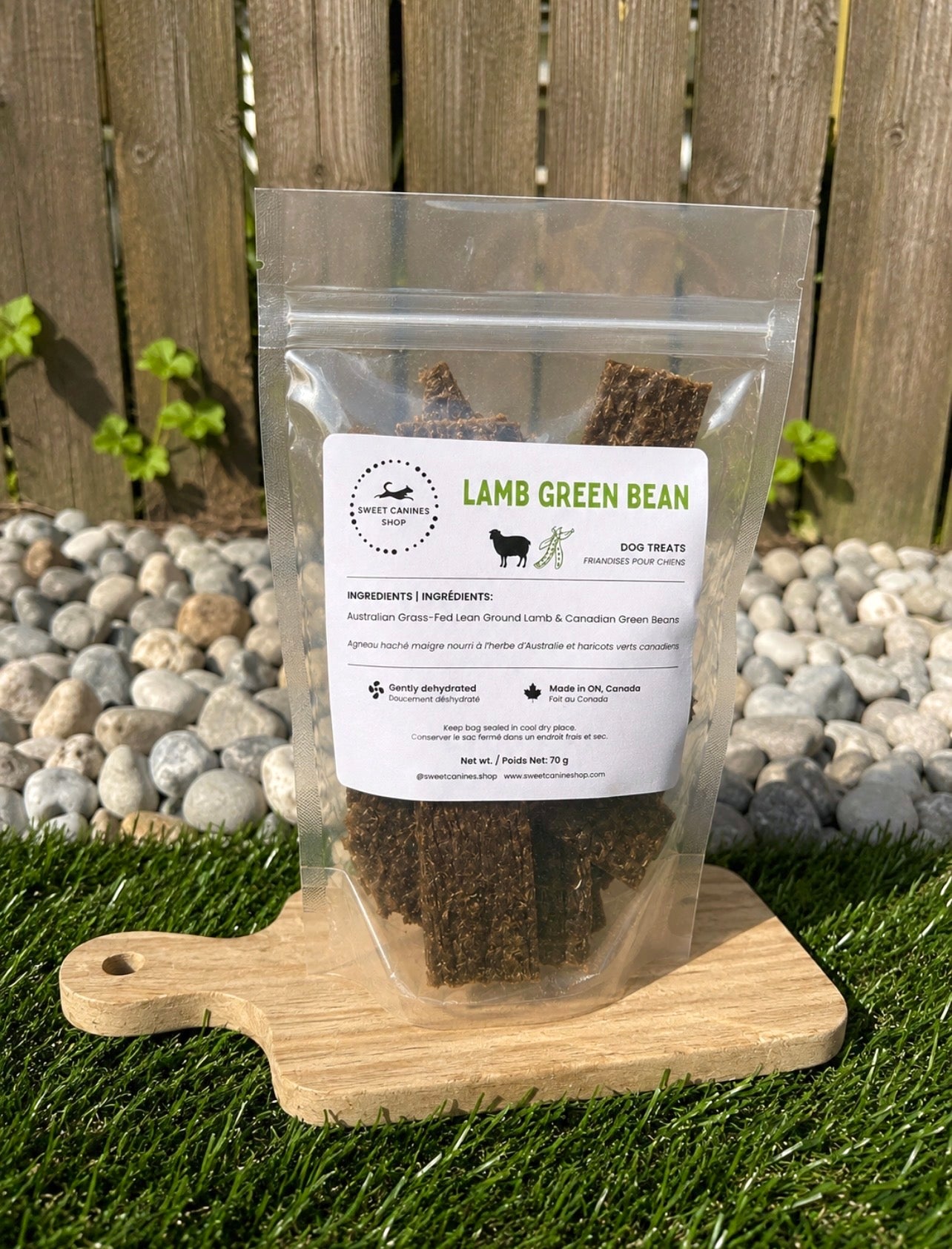 A clear stand-up pouch of dog treats labeled 'LAMB GREEN BEAN' by sweet canines shop placed on a wooden cutting board outdoors on grass with a fence and stones