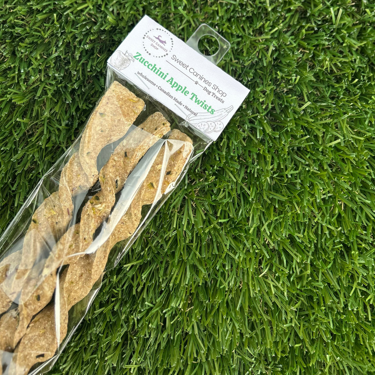 A packaged dog chew treat on artificial green grass. The clear plastic bag contains several tan, dried apple zucchini flavored dog chews, with a white paper header card attached.