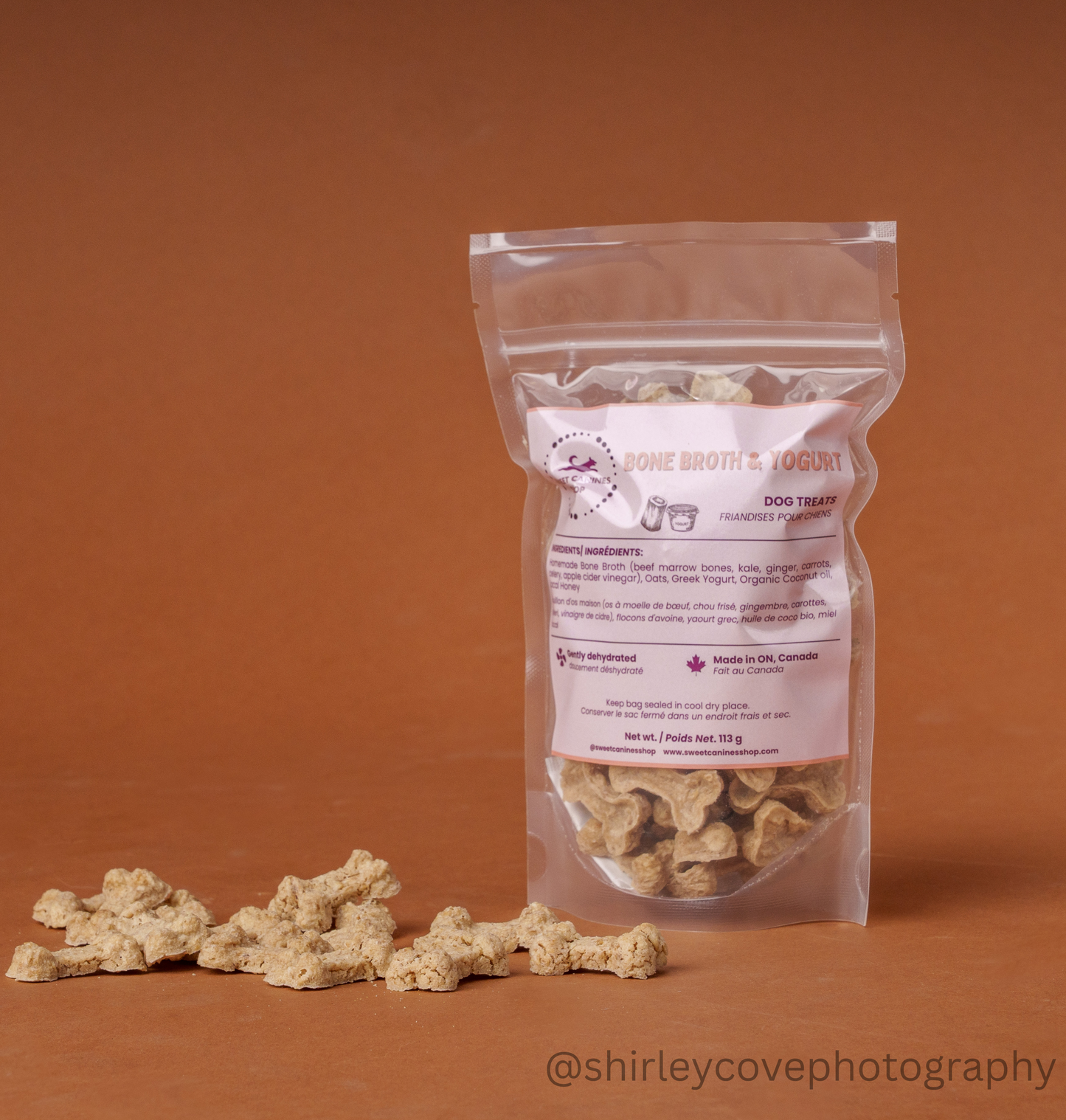 A clear stand-up pouch of dog treats stands on a warm brown studio background, with several bone-shaped treats scattered in front. The label reads 'Pone Broth & Yogurt' and 'Dog Treats.