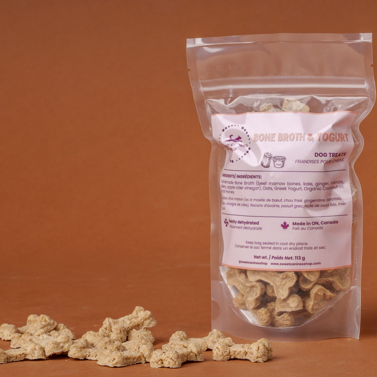 A clear stand-up pouch of dog treats stands on a warm brown studio background, with several bone-shaped treats scattered in front. The label reads 'bone Broth & Yogurt' and 'Dog Treats.