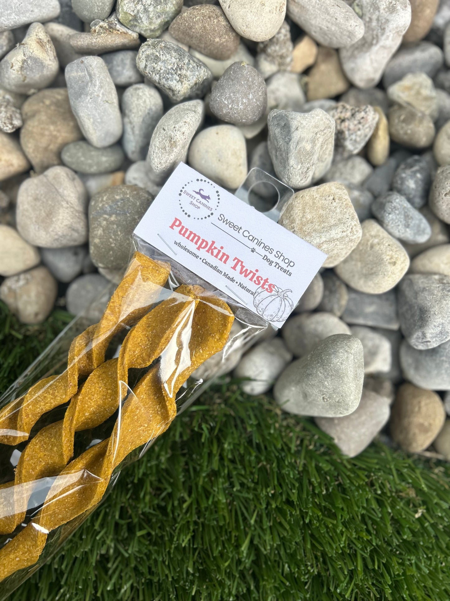 A packaged novelty dog treat toy shaped like twisted spaghetti, labeled 'Pumpkin Twists' by Sweet Canines Shop, placed on artificial green grass with a background of gray river stones.