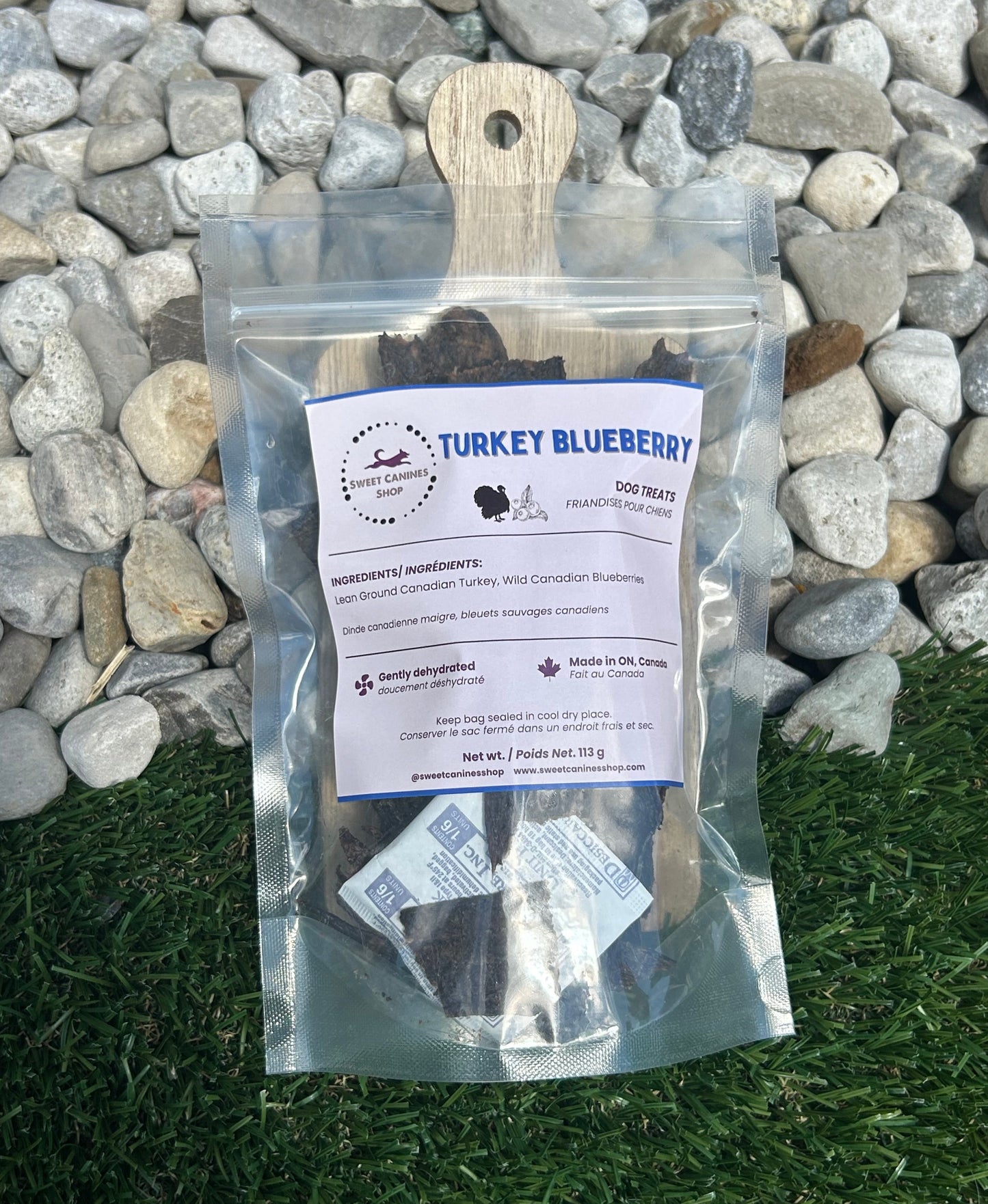 A clear resealable pouch of Turkey Blueberry dog treats sits on green grass in front of a bed of gray stones. The pouch has a wooden handle and a white label with blue text.