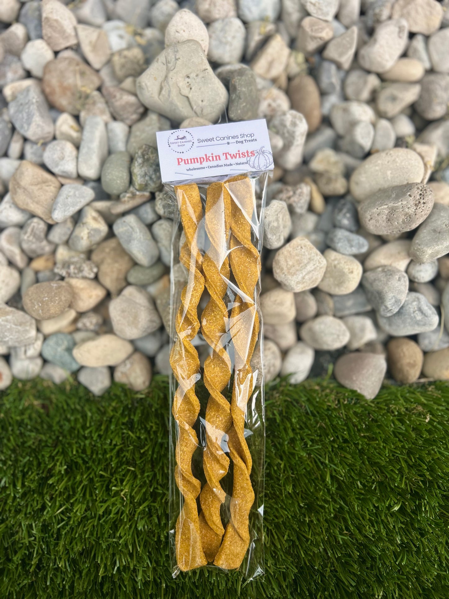 A package of natural pumpkin-shaped dog treats standing upright on artificial grass with a background of gray river stones. The treats are golden-brown and twisted, sealed in a clear plastic bag with a white paper label that reads 'Natural Pumpkin Twists' and 'Pumpkin Twists'.