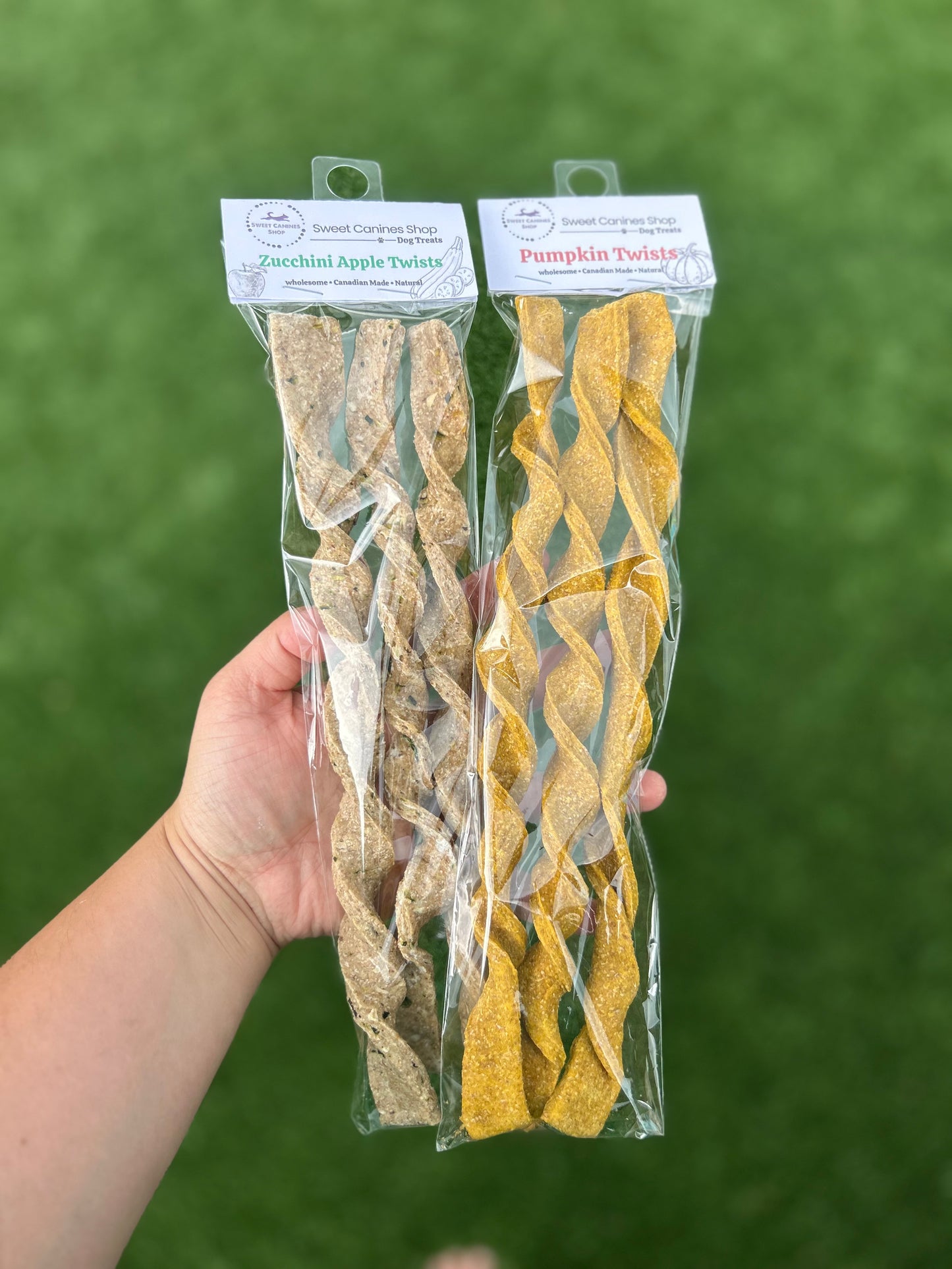A hand holds two packaged dog treats against a green grassy background. The left package contains zucchini apple twists in a light tan color, and the right package contains pumpkin twists in a golden yellow color.