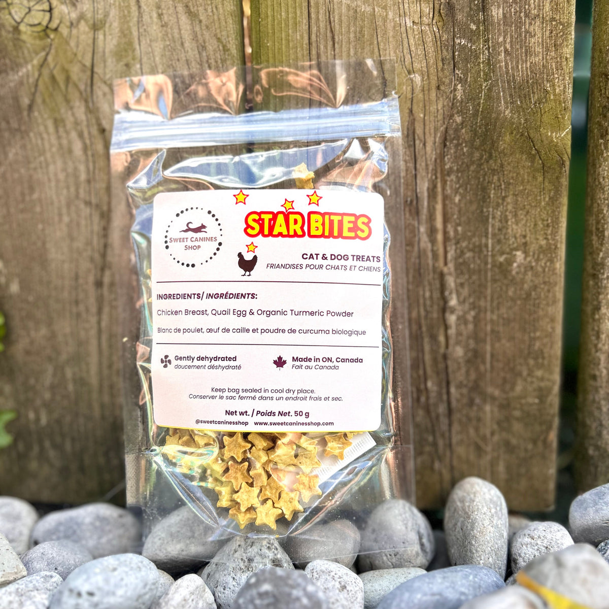 A clear plastic bag of Star Bites Cat & Dog Treats stands upright against a weathered wooden fence, with smooth gray stones in the foreground. The label shows the Star Bites logo and product details.