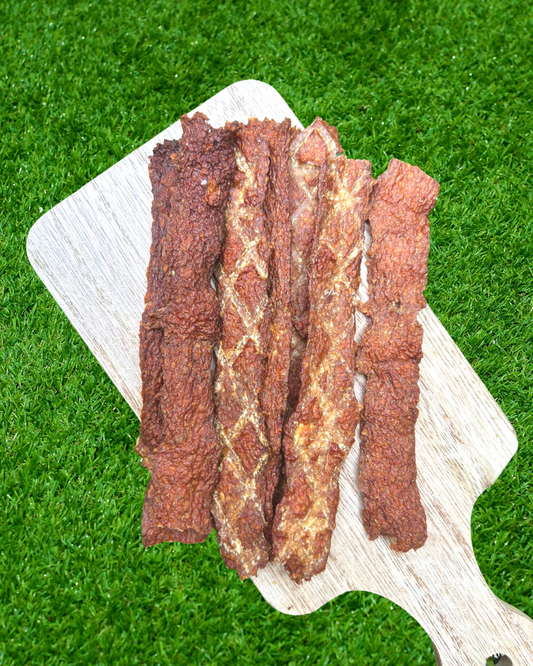 Four strips of textured, reddish-brown pork jerky arranged on a light-colored wooden cutting board, set against a bright green artificial grass background.