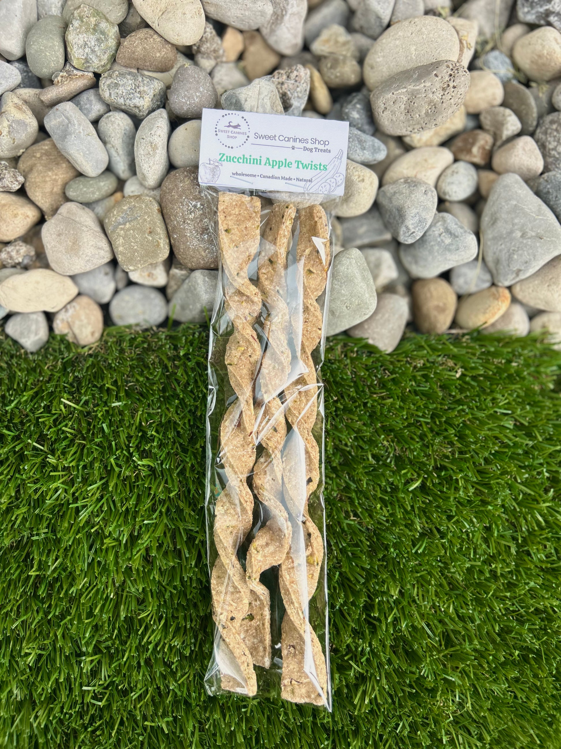 A package of spiral-shaped apple zucchini treats standing upright on artificial green grass with a bed of gray river stones behind it. The clear plastic bag contains several light tan, twisted pie crust sticks, and a white paper label at the top reads 'Sweet canines shop’ 