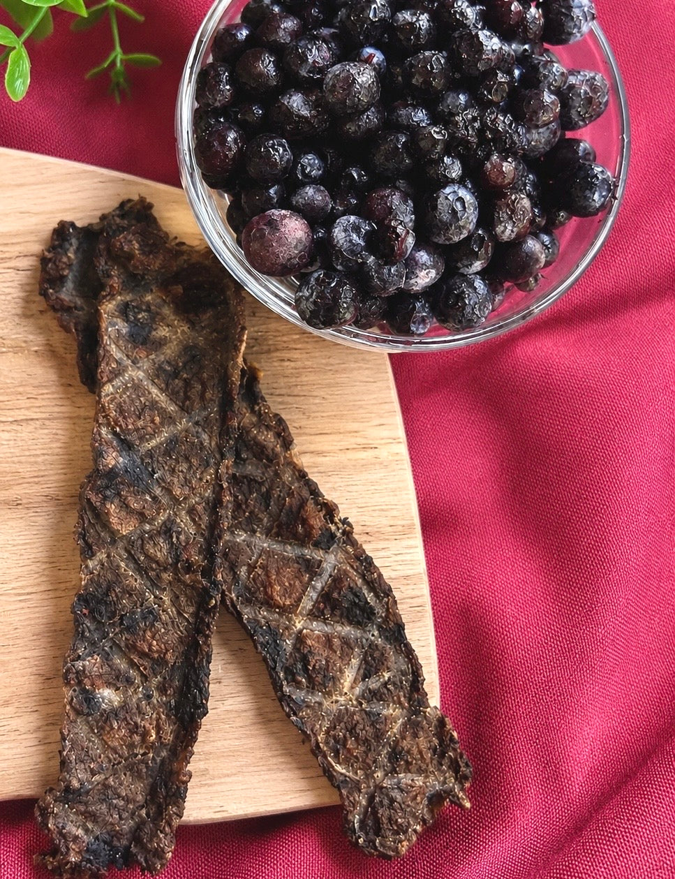 Two strips of dark, crispy blueberry and beef jerky with a diamond grill pattern, displayed on a wooden cutting board next to a glass bowl filled with fresh blueberries on a red fabric background.