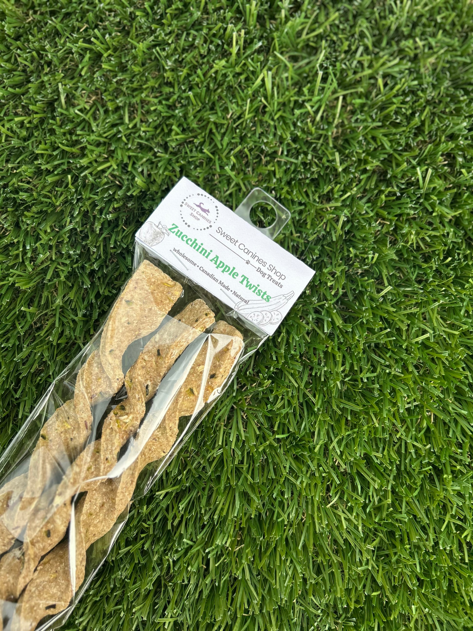 A packaged dog chew treat on artificial green grass. The clear plastic bag contains several tan, dried apple zucchini flavored dog chews, with a white paper header card attached.