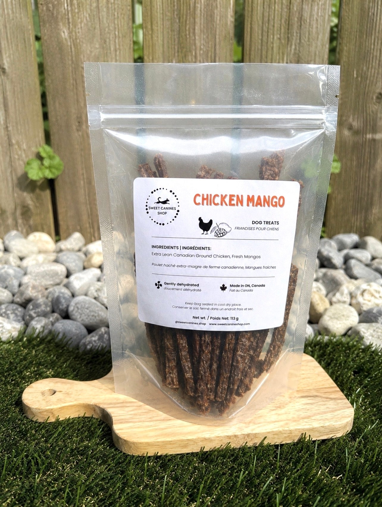 A clear stand-up pouch of dog treats labeled 'Chicken Mango' sits on a small wooden cutting board outdoors on grass, with a wooden fence and rocks behind it.