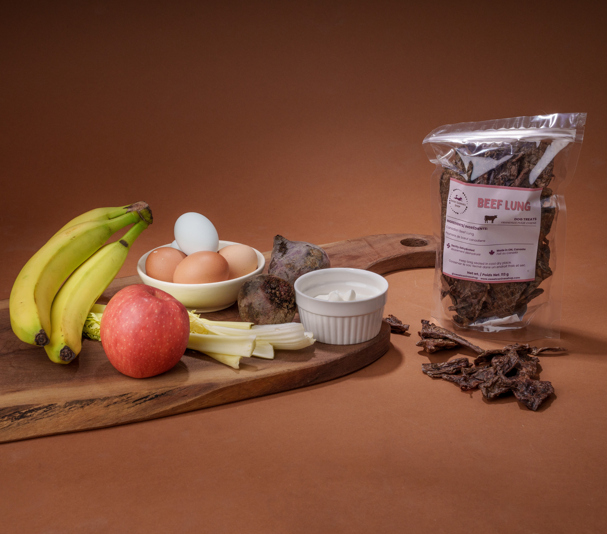 A clear stand-up pouch of beef jerky stands on a brown studio background next to a wooden cutting board with ingredients including bananas, an apple, eggs, celery, and a ramekin, with loose jerky pieces scattered beside it.
